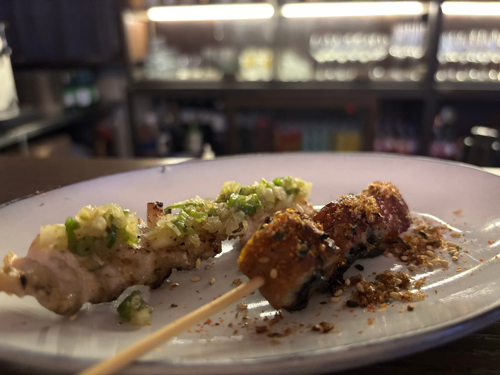 Two grilled skewers on a plate, with the bar and its glasses blurred behind